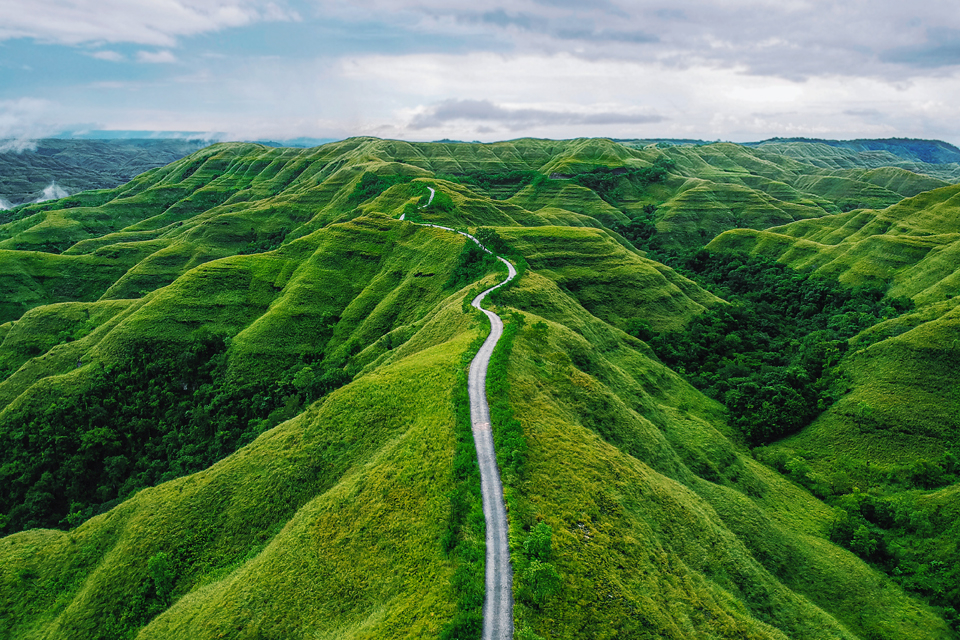 Hills, covered with vegetation, stretch toward the horizon, where clouds and sky are visible. At the center of the image, a winding road cuts through the hills heading in the direction of the horizon. 