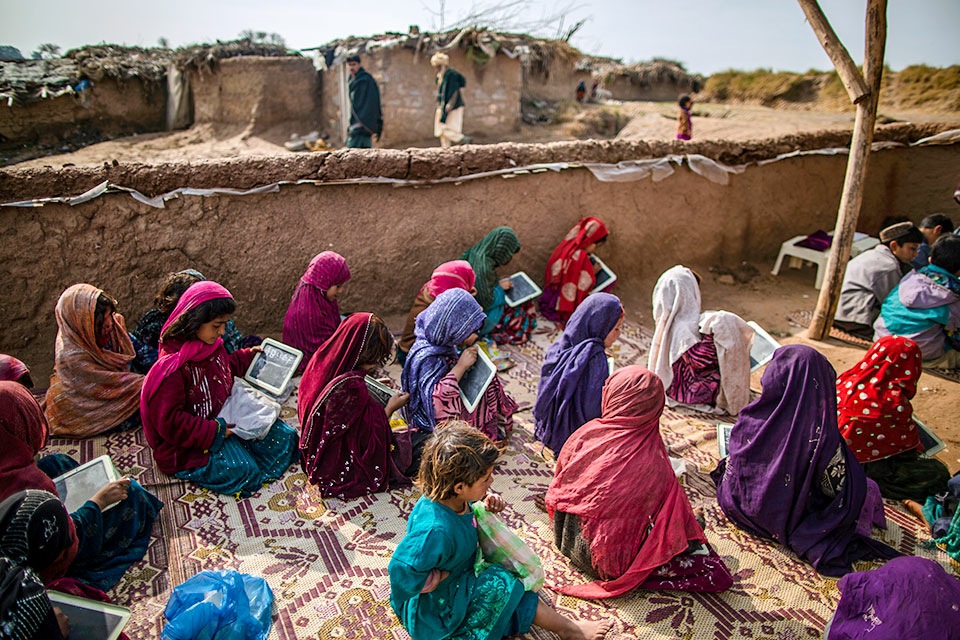 Afghan refugee girls in colorful headscarves seen from above as they kneel on blankets holding small personal chalk tablets while attending school in Islamabad, Pakistan.