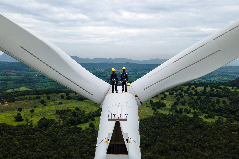 Two electrical engineers wearing protective gear stand between the blades at the top of a wind turbine in Thailand. Far below them, the land is densely forested. Another wind turbine sits in the distance and mountains are on the horizon.  
