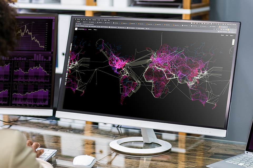 A person, with their back to the camera, sits at a desk in front of two computer monitors. The monitor on the right shows a world map that uses bright color to represent demographics, infrastructure, borders and resources. Similarly colored graphs are partially visible on the monitor to the left. 