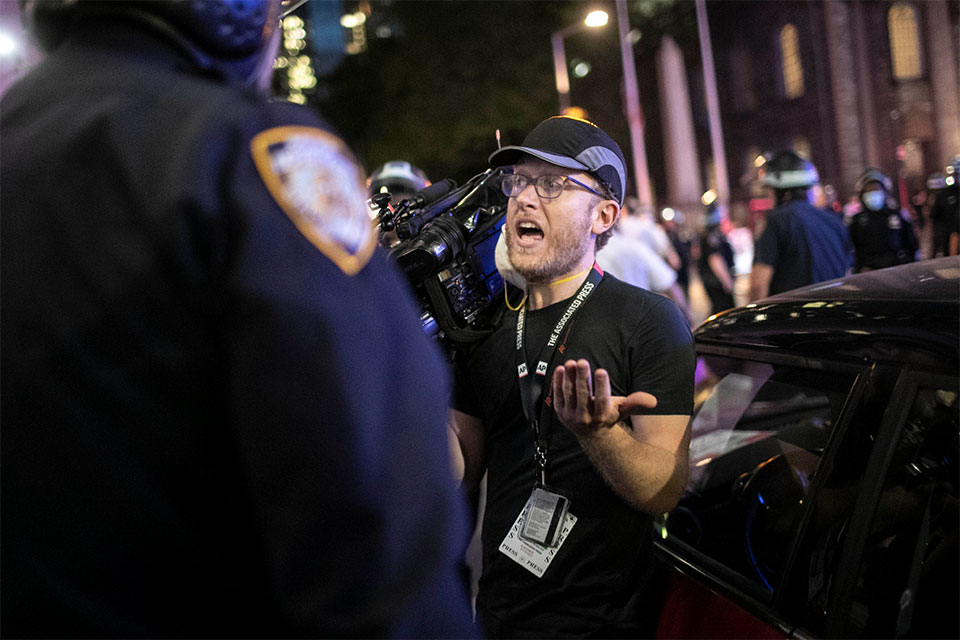 A journalist holding a TV camera on his right shoulder faces a police officer seen from behind while a protest is going on in the back.  The journalist is reminding the officer that the press are considered "essential workers" in regard to curfews related to protests.