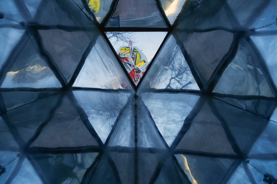 Triangular panels of glass create a wall. Light shows through some of the panels, making them appear brighter. Through a panel at the center, bright colors form an abstract pattern.