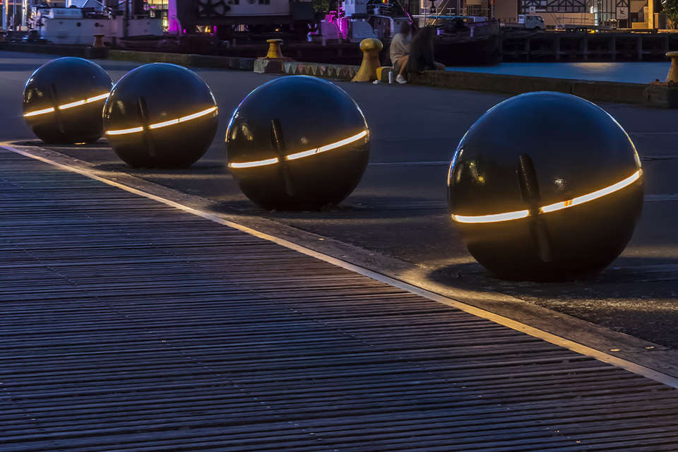 Four large metal spheres, each bisected by a strip of light, sit on the boardwalk along the harbor in Wellington, New Zealand. Water and the skyline are behind them. 