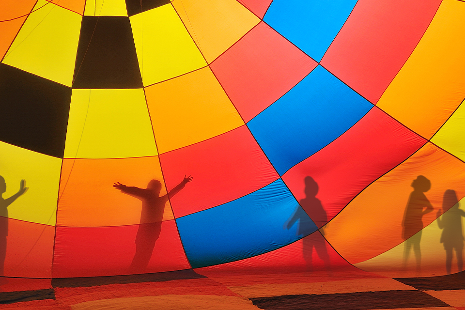A silhouetted view of five children playing outside a hot air balloon decorated in squares of red, yellow, orange, blue and black.
