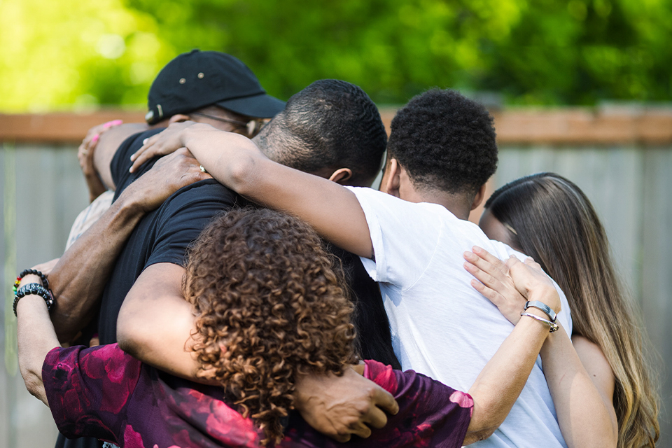 A group of five multi-generational family members, three male, two female, embrace in a backyard with their backs to the camera.