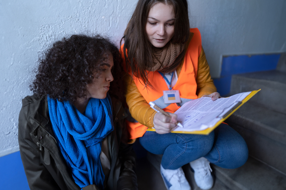 A female volunteer, right, and Ukrainian refugee woman, left, sit on steps in a train station, both looking at a document the volunteer is holding and explaining.