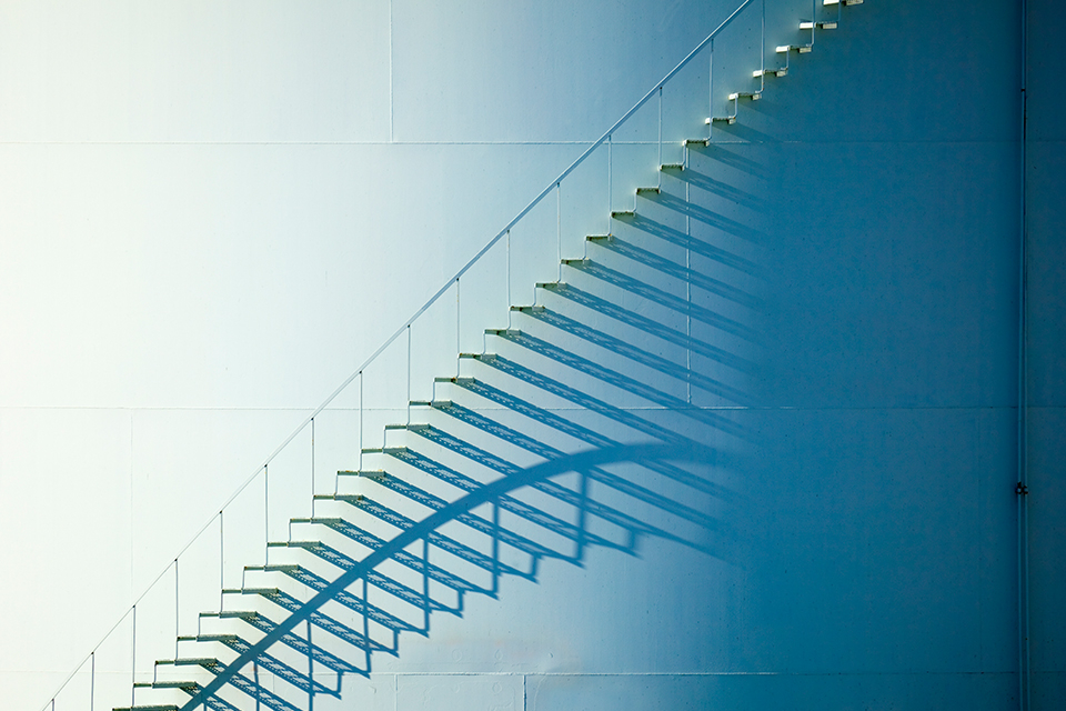 A white suspension staircase enveloped by a dark shadow on an oil storage tank. The shadow is located on the right side and covers about one third of the image.