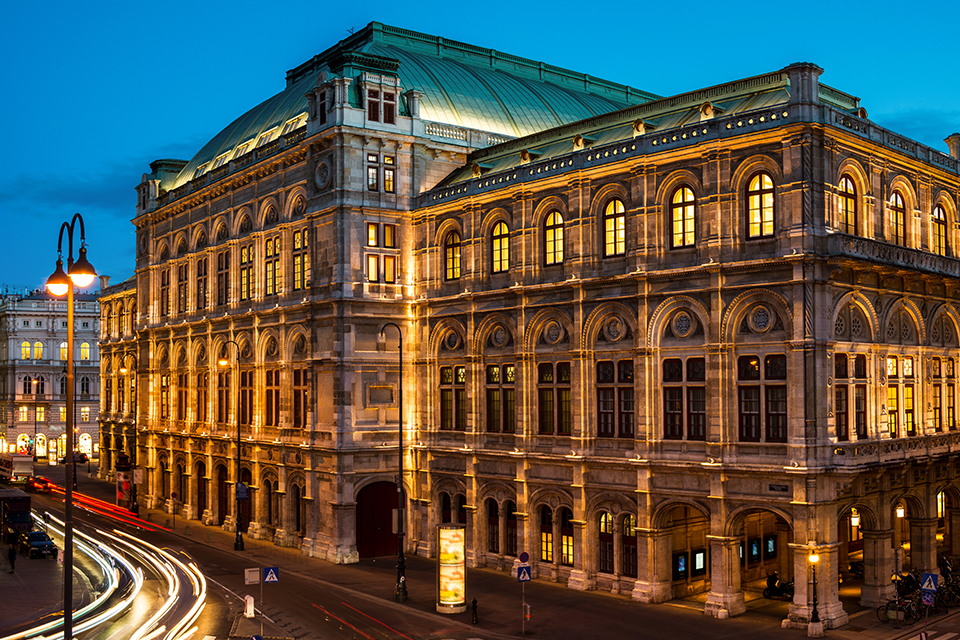 A nighttime view of the Wiener Staatsoper (Vienna State Opera) in Vienna, Austria. The Neo-Renaissance building is warmly lit, inside and out. Light from traffic appears as red and white streaks in the left foreground.
