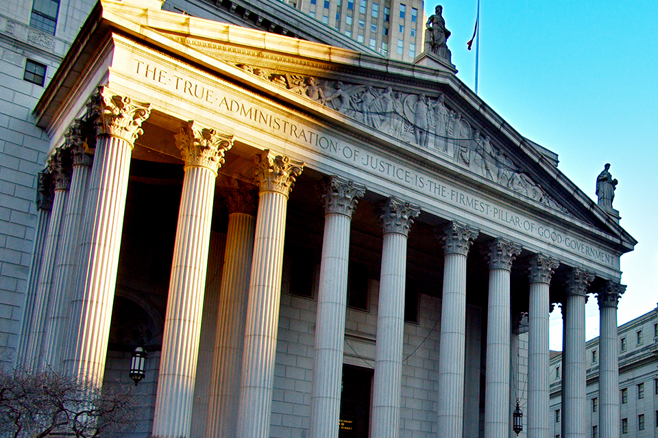 A view of the portico of the New York County Supreme Court, in lower Manhattan in New York City. Above its columns, the portico is engraved with the motto, "The true administration of justice is the firmest pillar of good government." Sun shines on the top left corner.