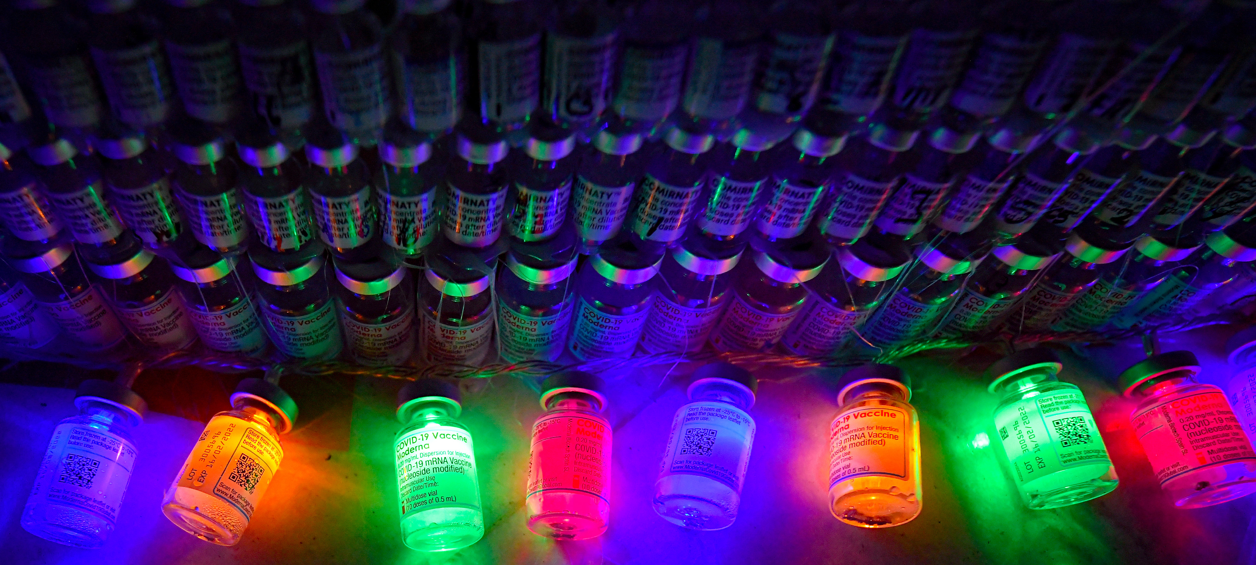 Bright, colorful electric lights inside empty COVID-19 vaccine containers shine beside more empty containers, which are stacked to resemble a Christmas tree 