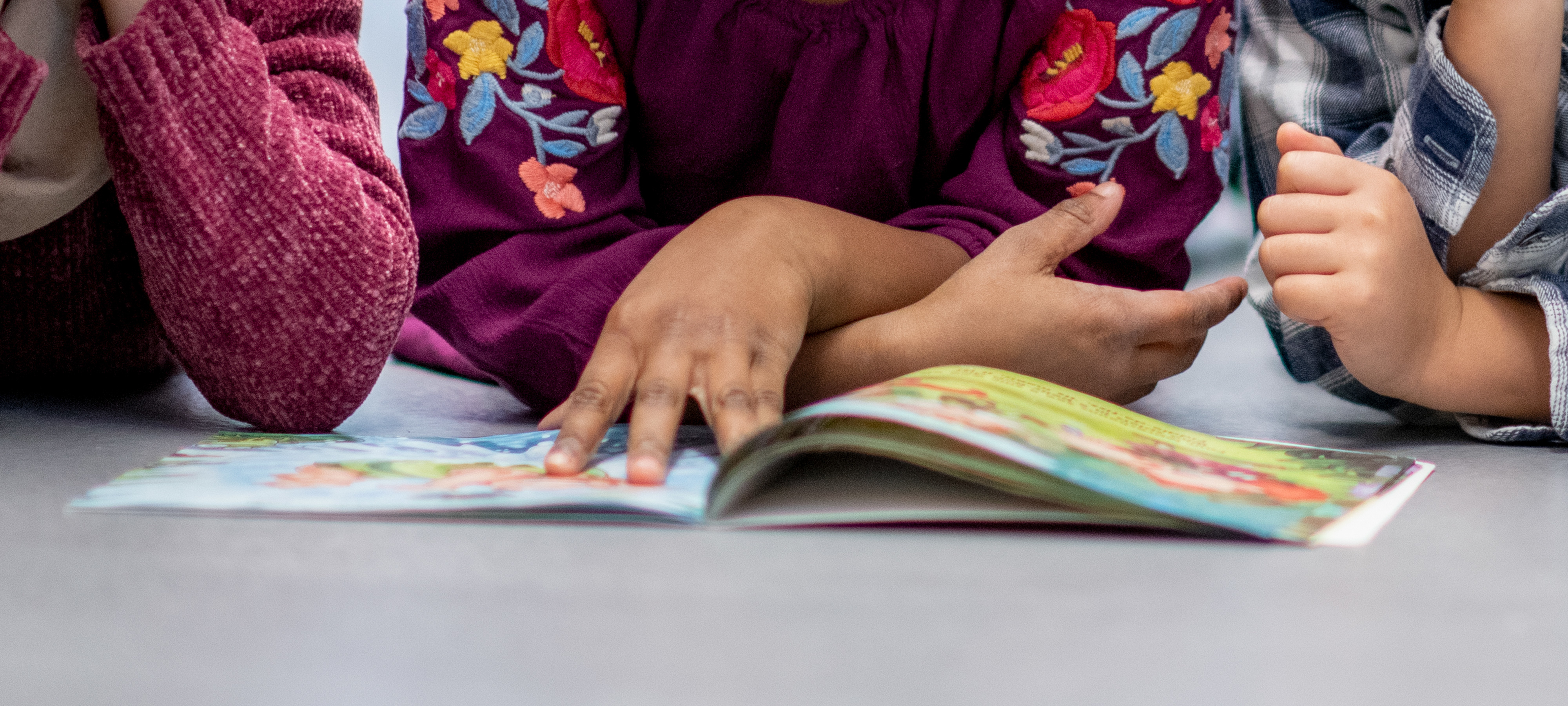 Close-up of the hands and arms of two young elementary school students who are lying together on the floor and reading a book that is on the floor in front of them.