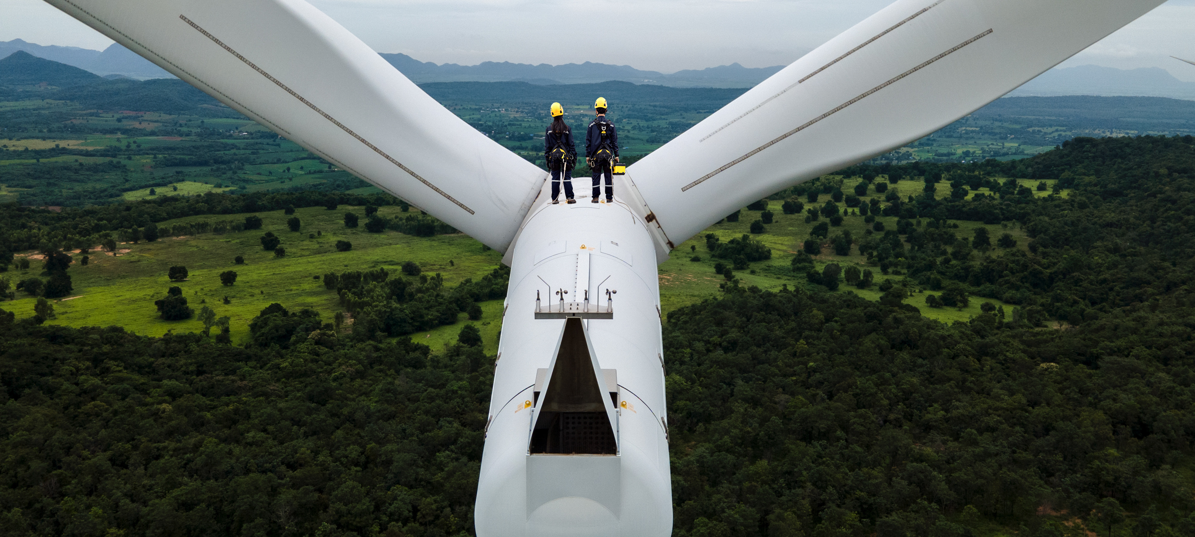 Two electrical engineers wearing protective gear stand between the blades at the top of a wind turbine in Thailand. Far below them, the land is densely forested. Another wind turbine sits in the distance and mountains are on the horizon.  