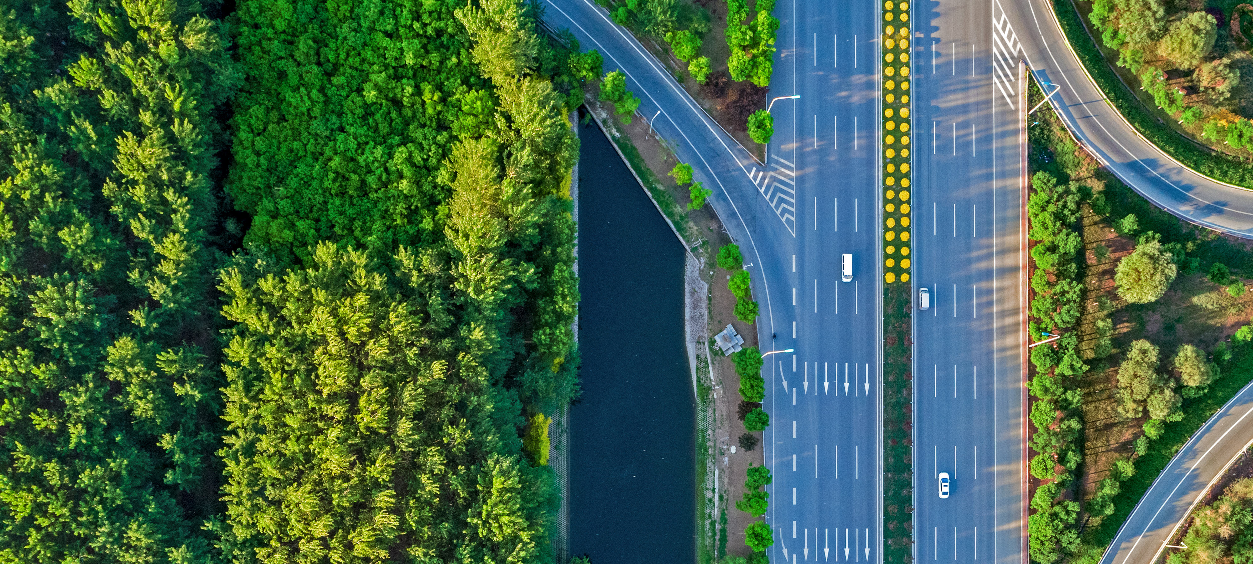 An aerial view of a mostly empty eight-lane highway in Beijing. A median divides the highway. One vehicle travels in one direction, and two travel in the other. Groves of trees surround the highway.