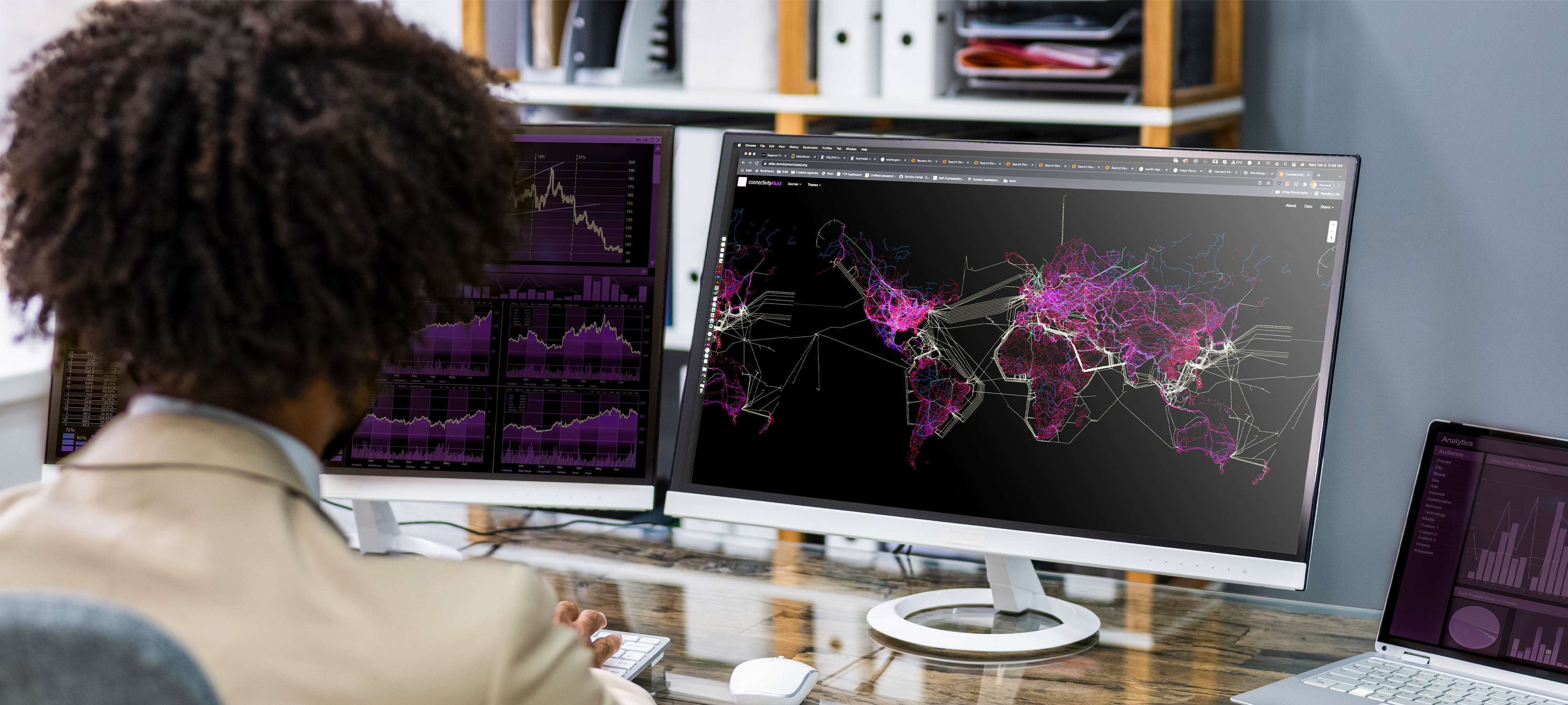 A person, with their back to the camera, sits at a desk in front of two computer monitors. The monitor on the right shows a world map that uses bright color to represent demographics, infrastructure, borders and resources. Similarly colored graphs are partially visible on the monitor to the left. 