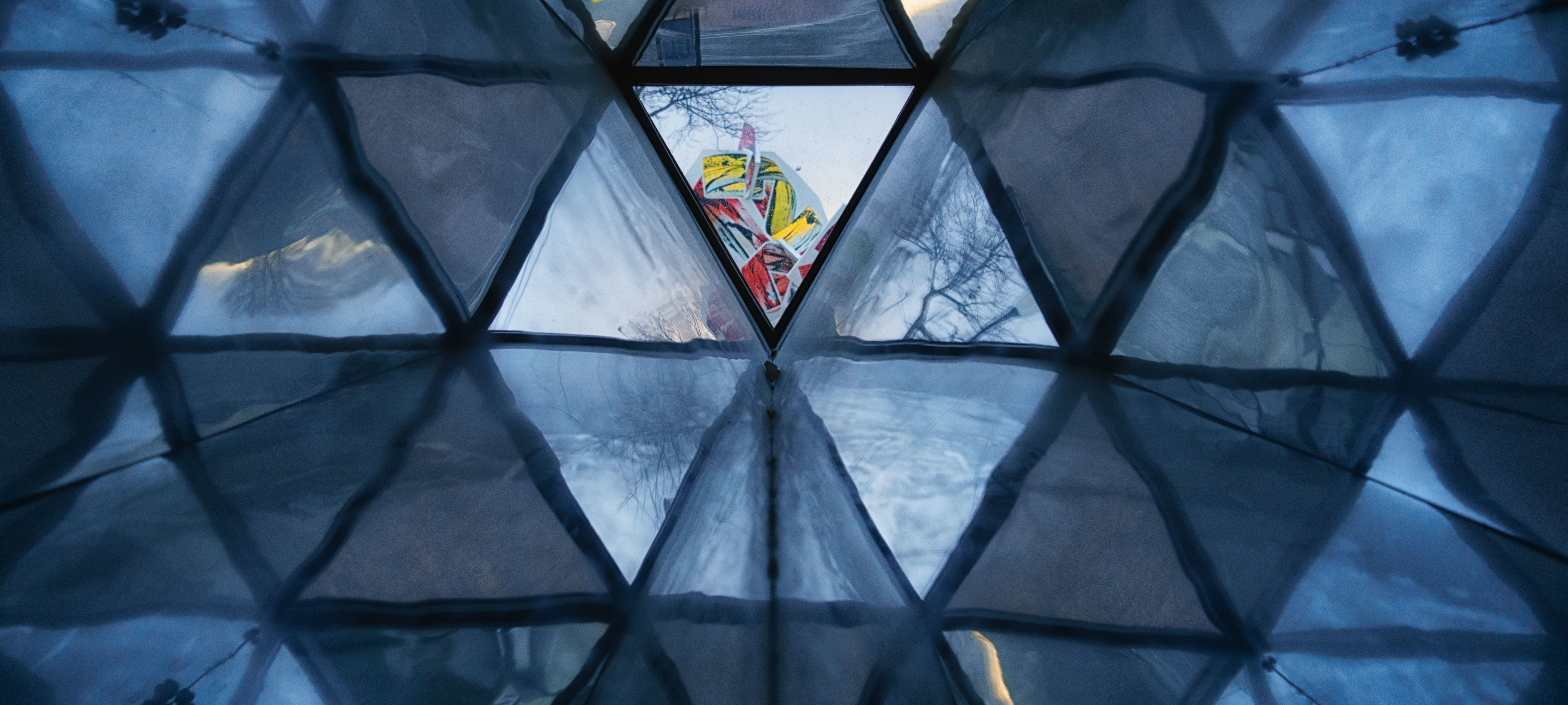 Triangular panels of glass create a wall. Light shows through some of the panels, making them appear brighter. Through a panel at the center, bright colors form an abstract pattern.