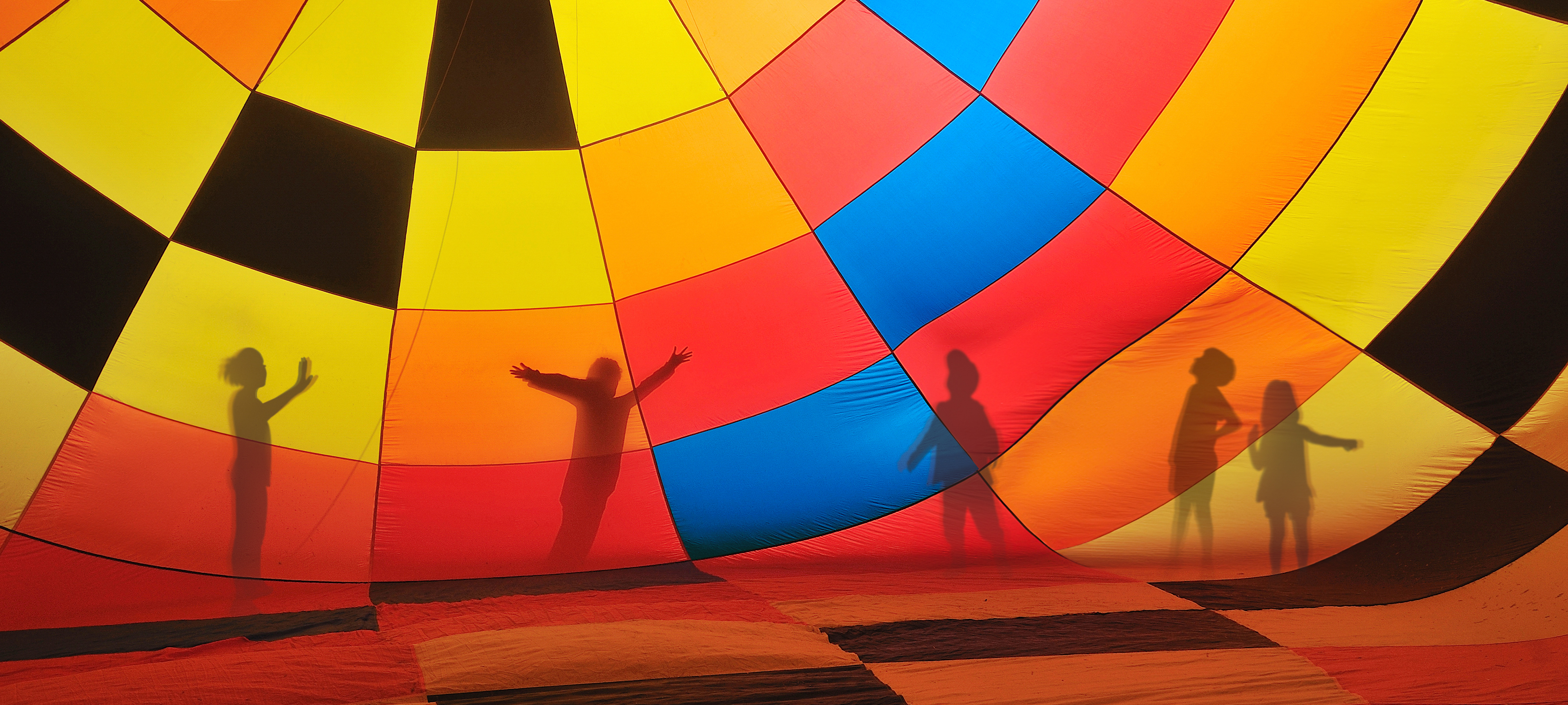 A silhouetted view of five children playing outside a hot air balloon decorated in squares of red, yellow, orange, blue and black.