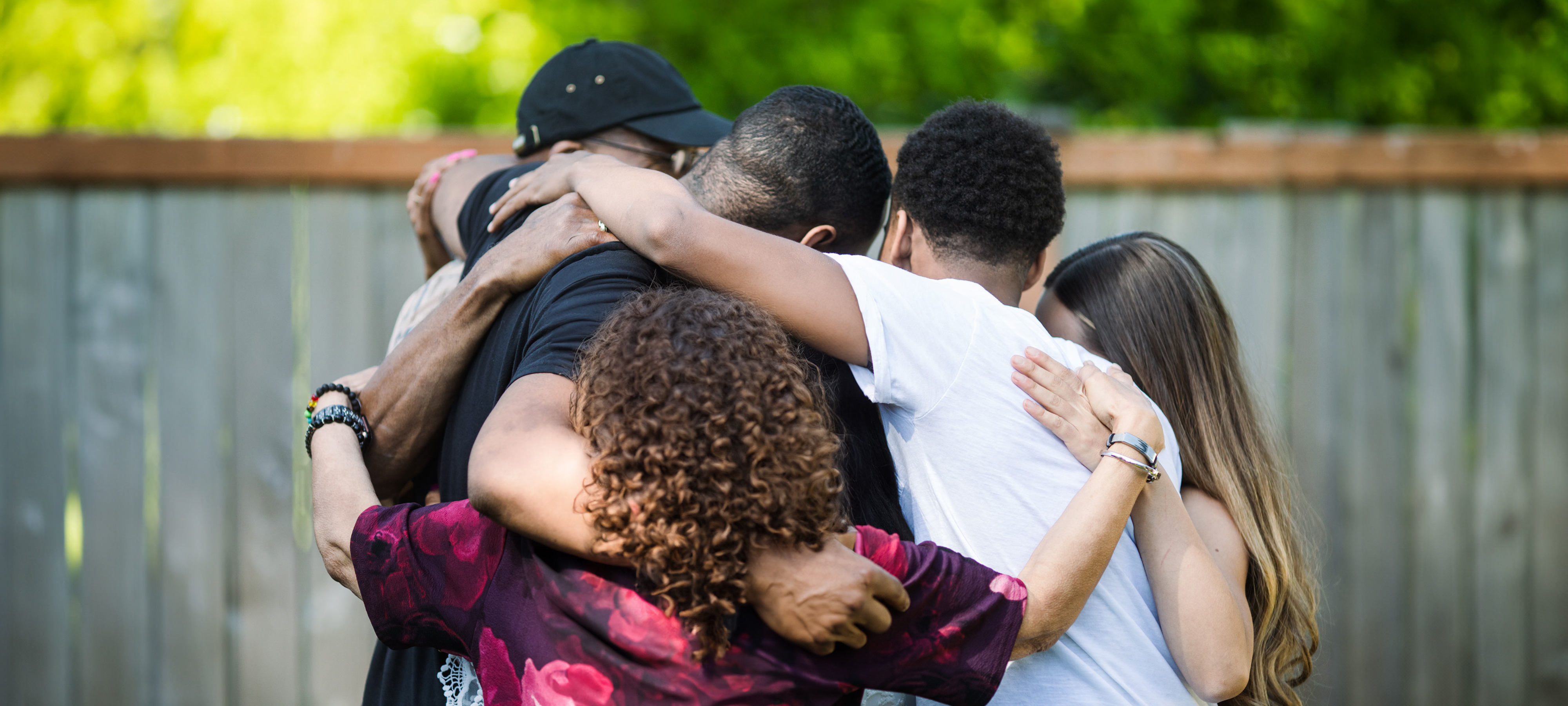 A group of five multi-generational family members, three male, two female, embrace in a backyard with their backs to the camera.