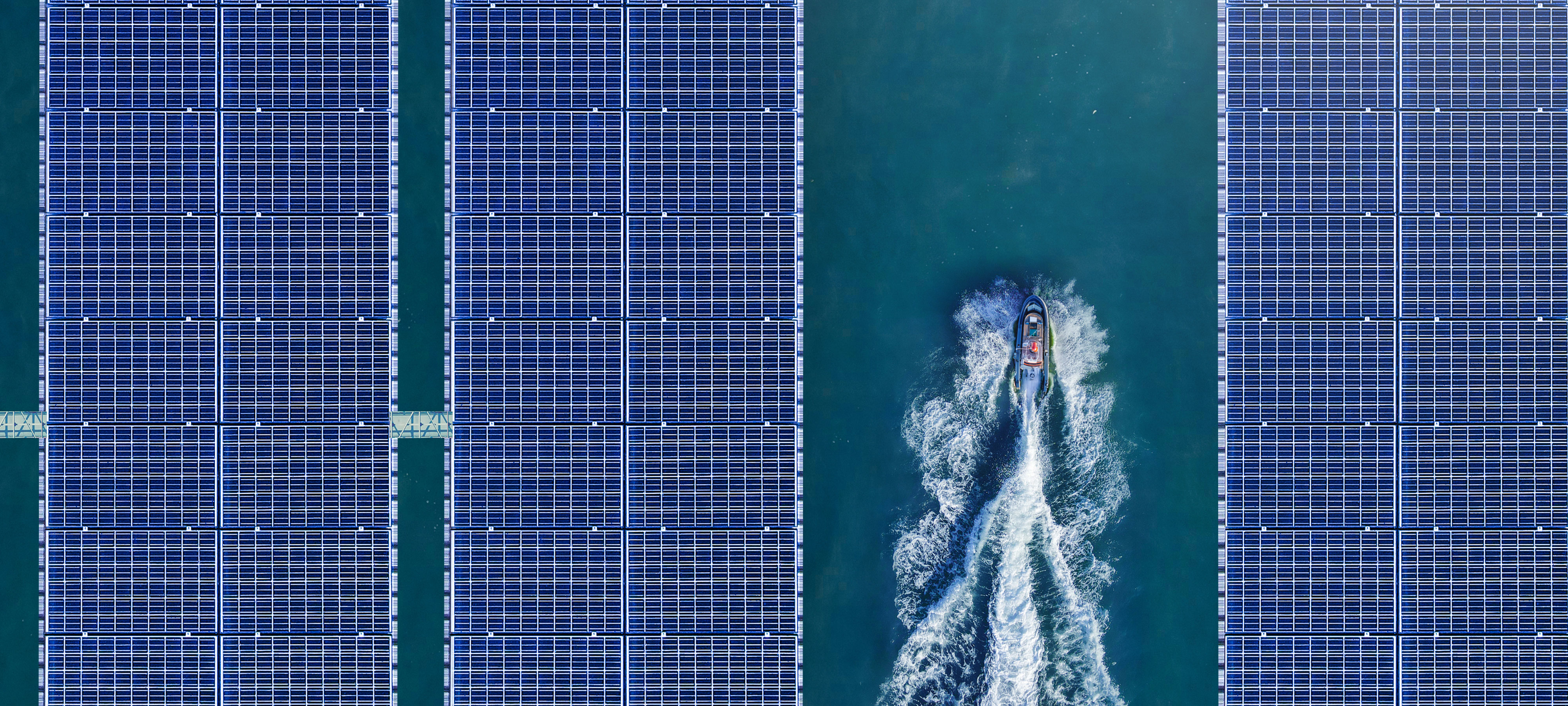 Bird’s-eye view of three solar panels floating in a dam, providing a source of renewable energy. There are a couple of people riding a jet ski between the two solar panels on the left side and the one on the right.