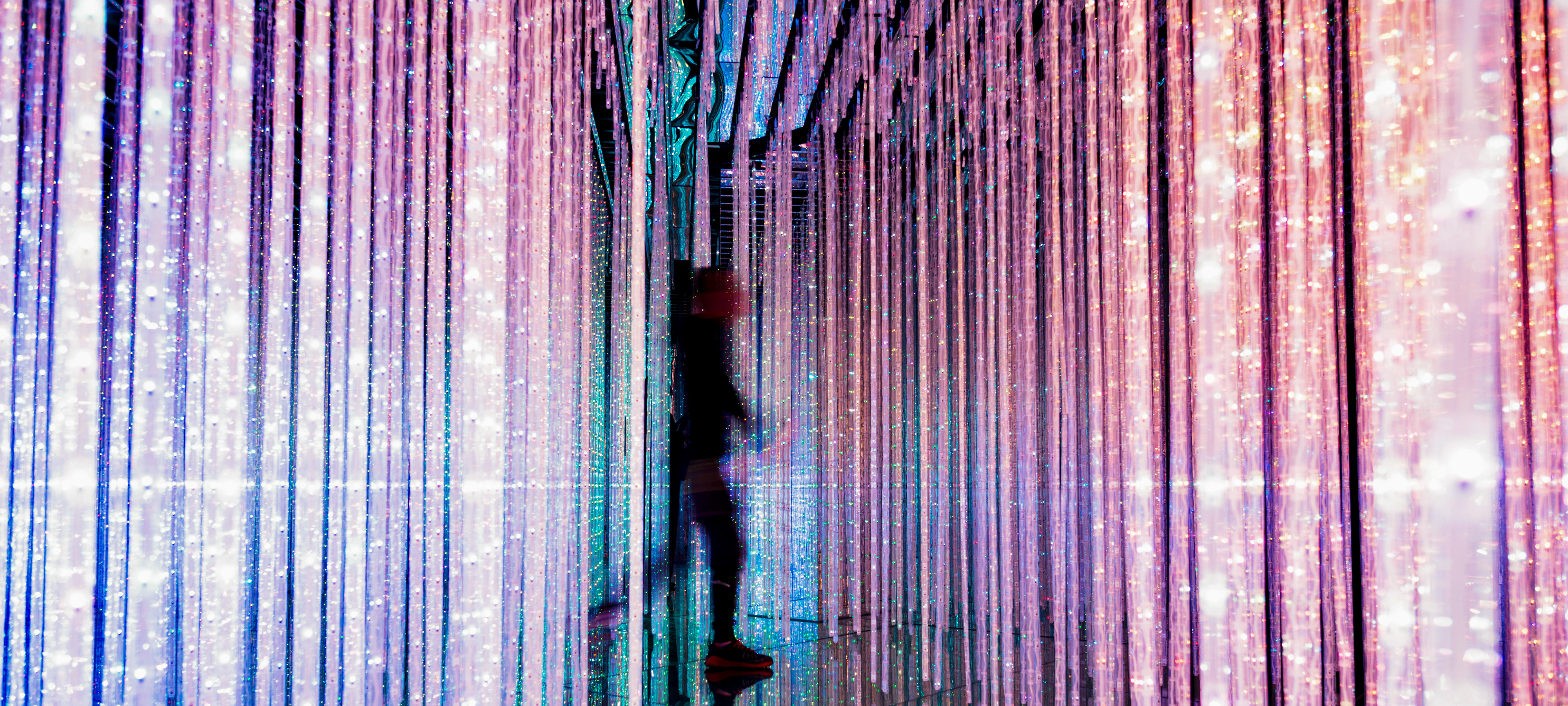 A blurry male figure walks through colorful strips of bright, sparkling lights. The lights cascade from the ceiling to the floor.