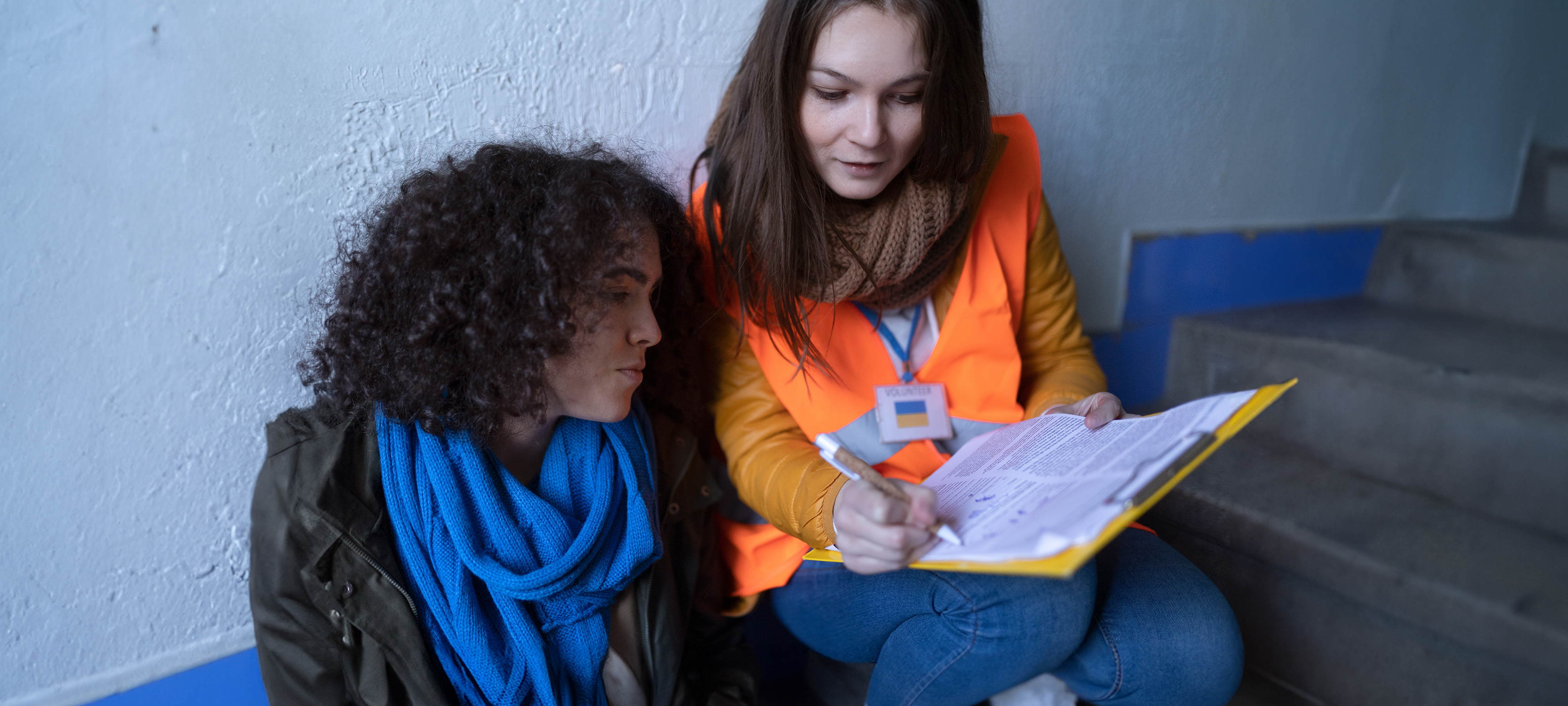 A female volunteer, right, and Ukrainian refugee woman, left, sit on steps in a train station, both looking at a document the volunteer is holding and explaining.