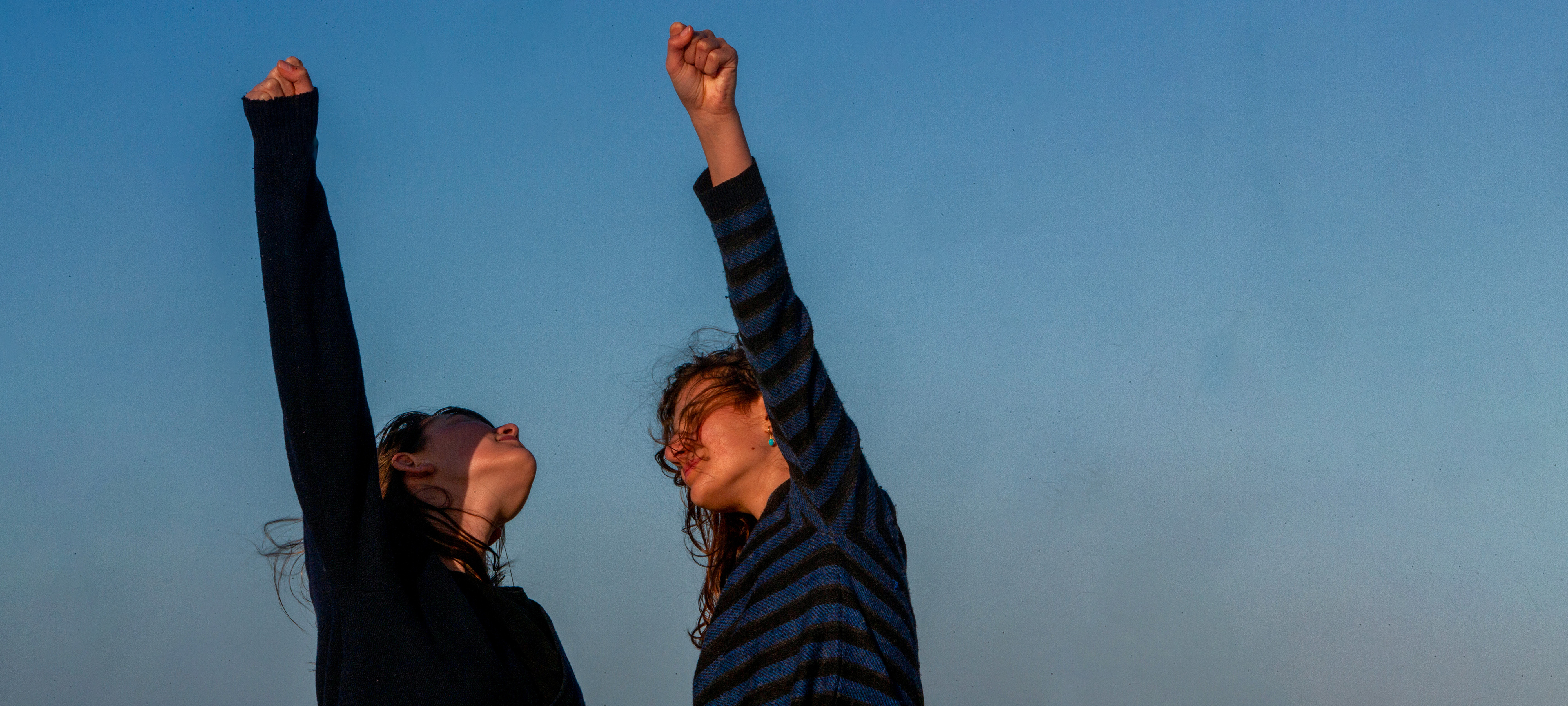 Two teenage girls facing each other raise their fists against a late-afternoon blue sky. One girl, left, looks up, while the other, right, looks into the distance. The wind blows their hair.