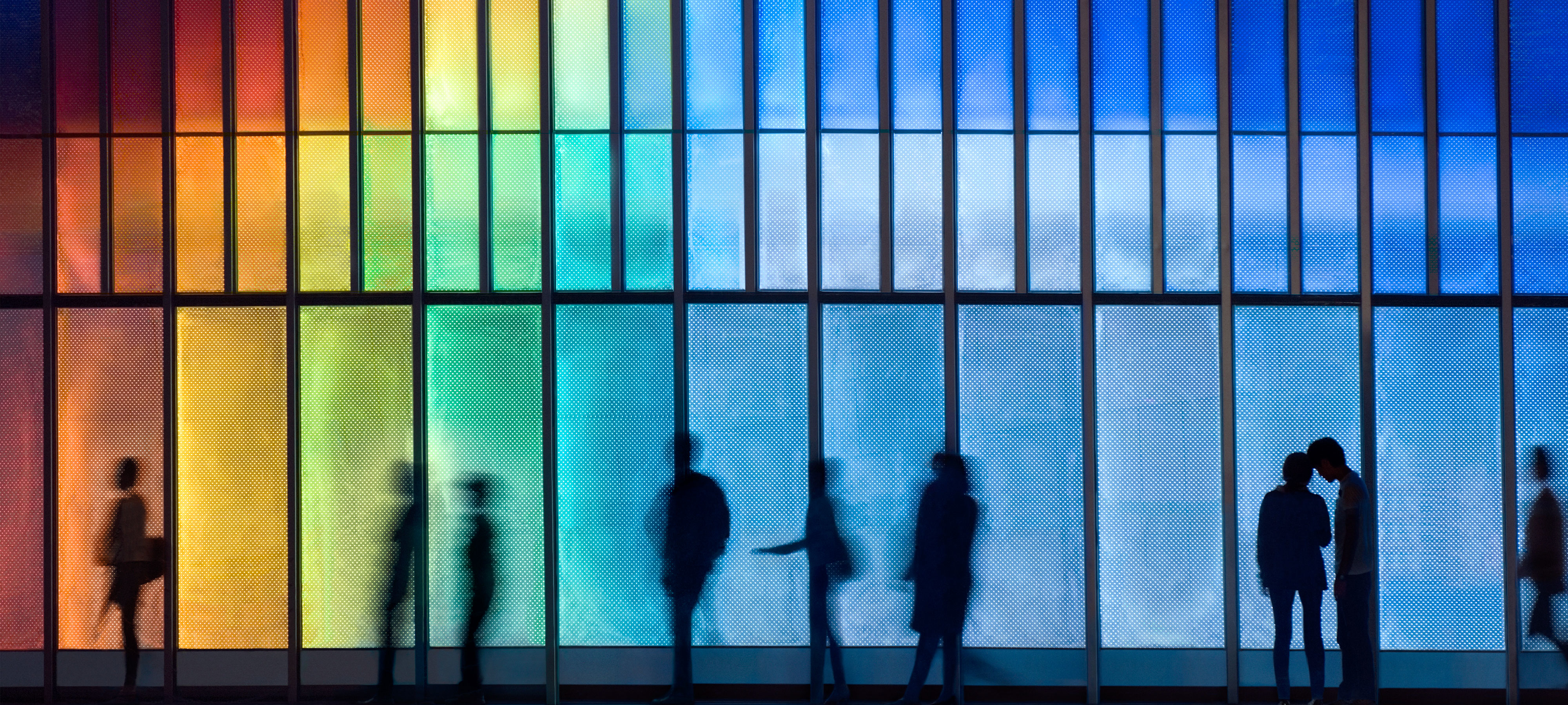 Silhouettes of individuals walking past an LED-lit façade of a building in Hong Kong, China, showcasing vibrant colors that constantly change. There are also two people talking in front of the building.