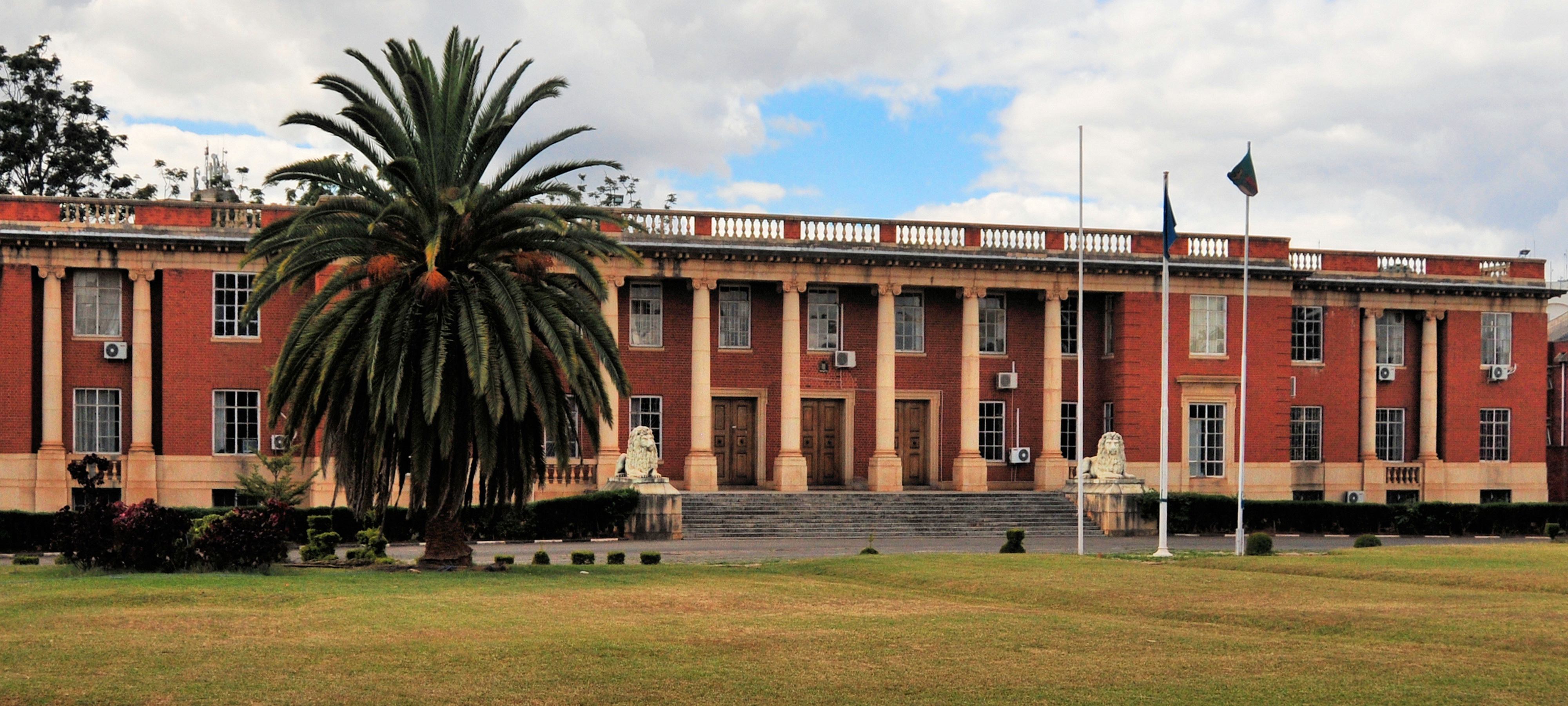 A distant view of the Supreme Court of Zambia, in the country's capital of Lusaka. The red-brick, two-story building features white columns along the main facade and a pair of white lion sculptures enclosing the wide entrance steps. A large palm tree stands to the left, and three flagpoles to the right.