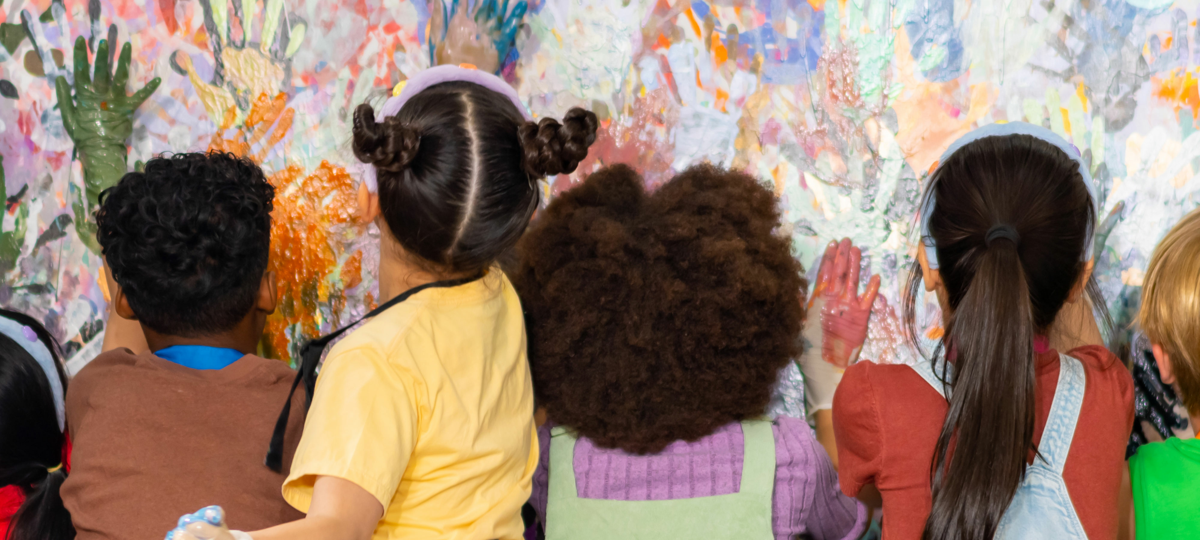 Six primary school-aged boys and girls from diverse backgrounds use their handprints to create a colorful abstract painting in a classroom. The children are different heights and are wearing colorful clothing.