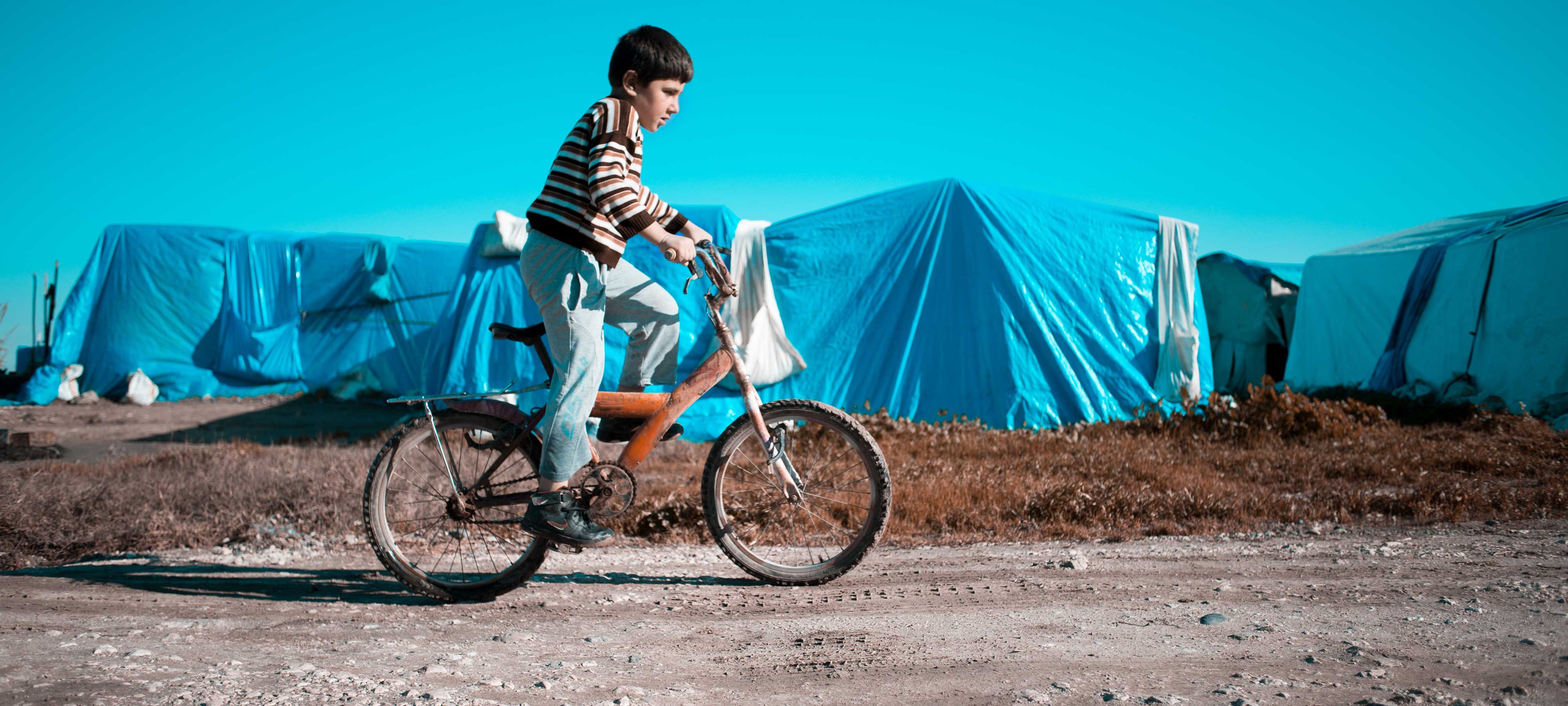 A child in a striped shirt and light-colored pants rides a bicycle along a dirt road during the day, passing in front of a refugee camp. The camp consists of several blue tents.