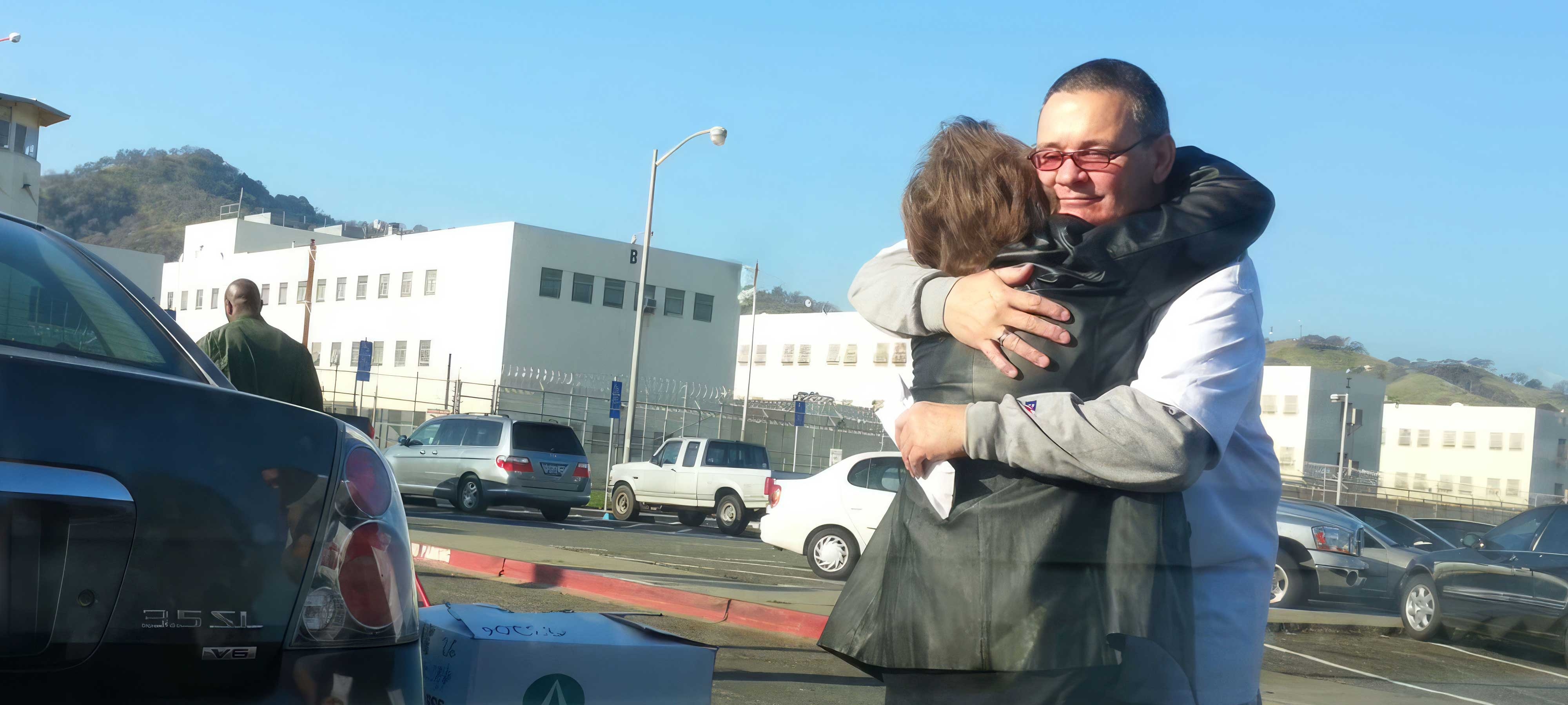 Two people, one n wearing glasses and a short-sleeve shirt over a long-sleeve shirt, embrace in a parking lot outside a prison during the day. The lot, separated from the white prison by a fence, contains several cars and SUVs.